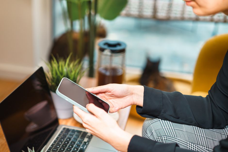 Mulher trabalhando remotamente de casa, usando um smartphone e um laptop em uma mesa aconchegante com uma planta e um gato ao fundo.