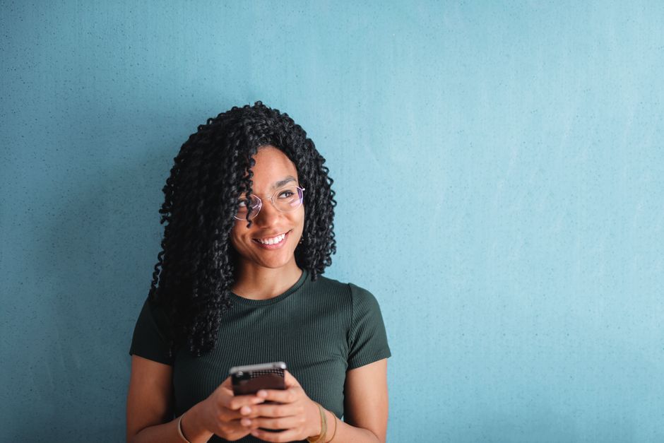 Uma jovem alegre com cabelo encaracolado usa seu celular contra um fundo azul.