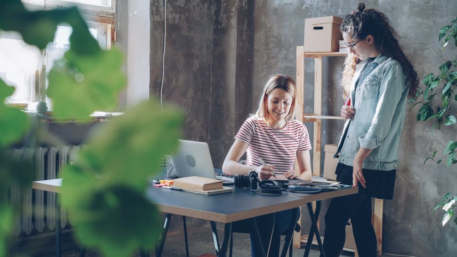 Duas mulheres trabalhando juntas em uma mesa em um escritório moderno recheado de plantas.