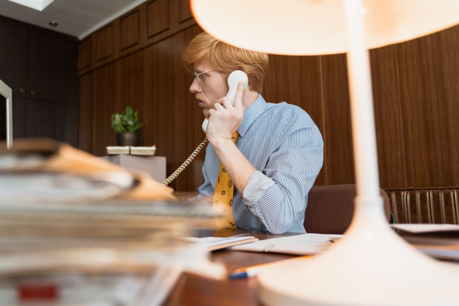 Homem profissional com óculos falando em um telefone vintage em um ambiente de escritório.