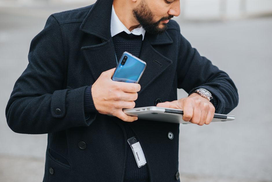Um homem profissional ao ar livre verificando a hora enquanto segura um smartphone e um laptop.