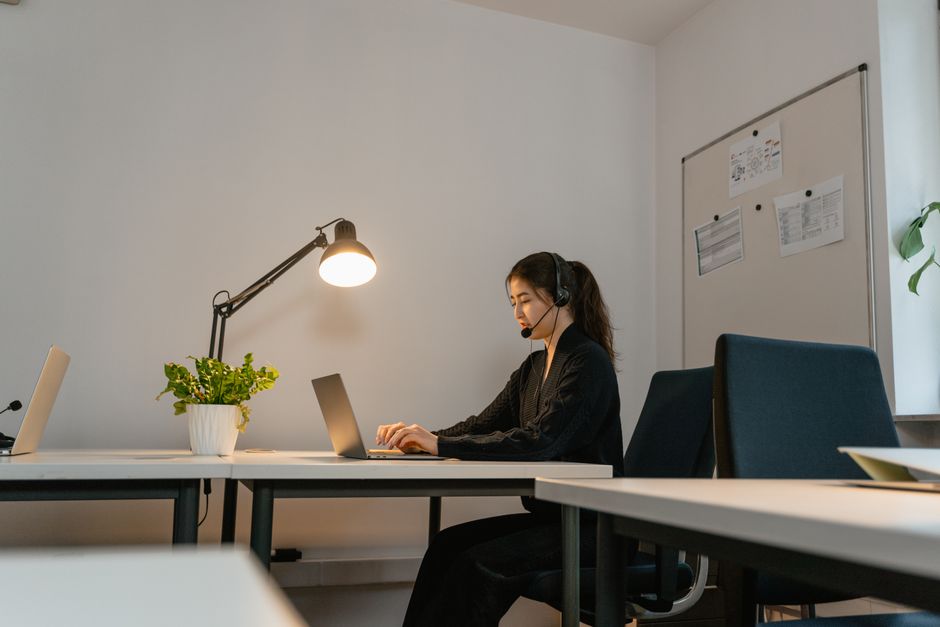 Uma mulher em um ambiente de escritório moderno, usando um fone de ouvido e trabalhando em um laptop sob uma luminária de mesa.