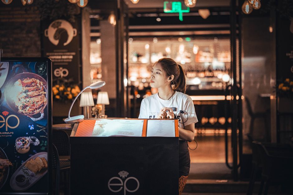 Uma jovem recepcionista está na entrada de um restaurante moderno, pronta para receber os clientes.
